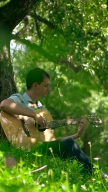 Dikey. Yapraklı bir gölgelik altında akustik gitar çalan bir adam, benekli güneş ışığıyla yıkanmış. nazik tıngırtı, yemyeşil, ve huzurlu bir melodi. Akustik gitar müziği orman havasını doldurur, bir adamın parmakları tellerde dans eder.. 