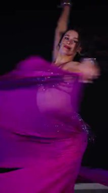 Dikey. Zarif giyimli bir kadın, müziğe gidiyor ve karanlık bir ortamda dans ediyor. Mor elbiseli kadın loş ışıklı odada zarif bir şekilde göbek dansı yapıyor. Gölgeli alanda mor elbiseler giyen kadın dansçı. Karanlıktaki kadın 