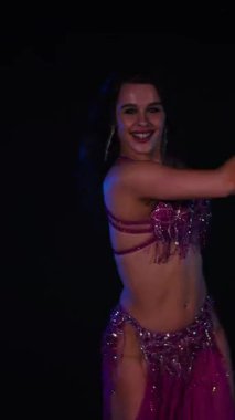 Dikey. Zarif giyimli bir kadın, müziğe gidiyor ve karanlık bir ortamda dans ediyor. Mor elbiseli kadın loş ışıklı odada zarif bir şekilde göbek dansı yapıyor. Gölgeli alanda mor elbiseler giyen kadın dansçı. Karanlıktaki kadın 