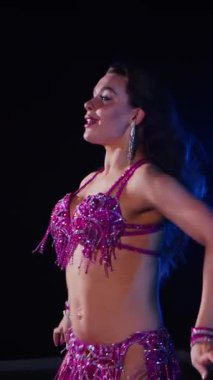 Dikey. Zarif giyimli bir kadın, müziğe gidiyor ve karanlık bir ortamda dans ediyor. Mor elbiseli kadın loş ışıklı odada zarif bir şekilde göbek dansı yapıyor. Gölgeli alanda mor elbiseler giyen kadın dansçı. Karanlıktaki kadın 