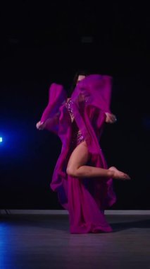 Dikey. Mor elbiseli kadın loş ışıklı odada zarif bir şekilde göbek dansı yapıyor. Yavaş çekim. Zarif giyimli bir kadın, müziğe gidiyor ve karanlık bir ortamda dans ediyor. Mor elbiseler giyen kadın dansçı Gölgeli alanda girdaplar.