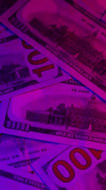 Dikey. Canlı bir Amerikan para akışı var. Çok renkli ışıkta yıkanmış bir Amerikan doları yağmuru. ABD banknotlarının yakın plan görüntüsü. Detayları renkli bir renge bürünmüş. kağıt para döner.
