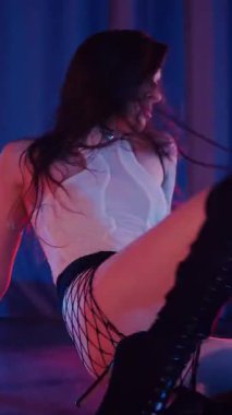 Dikey. Külotlu, tişörtlü, siyah çizmeli, baştan çıkarıcı dans eden seksi bir kadın. Bir dansçı kız karanlık bir salonda erotik bir şekilde dans eder. Kıvırma, striptiz. Atmosferik renkli ışık, kulüp, sahne.