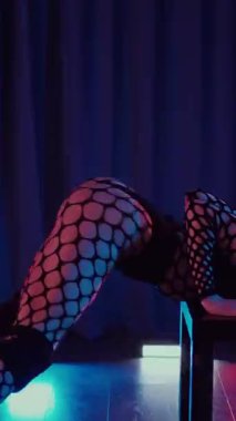 Dikey. Dans eden örgü giysili seksi kadın. Bir dansçı kız karanlık bir salonda erotik bir şekilde dans eder. Kıvranma, yüksek topuklar, striptiz, baştan çıkarıcı bir dans. Atmosferik renkli ışık, kulüp, sahne.
