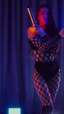 Dikey. Dans eden örgü giysili seksi kadın. Bir dansçı kız karanlık bir salonda erotik bir şekilde dans eder. Kıvranma, yüksek topuklar, striptiz, baştan çıkarıcı bir dans. Atmosferik renkli ışık, kulüp, sahne.