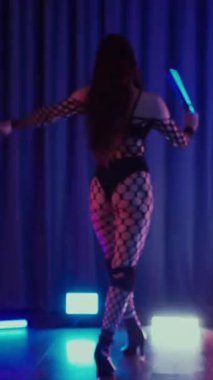 Dikey. Dans eden örgü giysili seksi kadın. Bir dansçı kız karanlık bir salonda erotik bir şekilde dans eder. Kıvranma, yüksek topuklar, striptiz, baştan çıkarıcı bir dans. Atmosferik renkli ışık, kulüp, sahne.