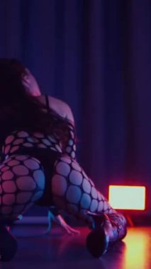 Dikey. Dans eden örgü giysili seksi kadın. Bir dansçı kız karanlık bir salonda erotik bir şekilde dans eder. Kıvranma, yüksek topuklar, striptiz, baştan çıkarıcı bir dans. Atmosferik renkli ışık, kulüp, sahne.