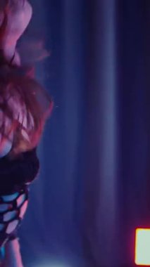 Dikey. Dans eden örgü giysili seksi kadın. Bir dansçı kız karanlık bir salonda erotik bir şekilde dans eder. Kıvranma, yüksek topuklar, striptiz, baştan çıkarıcı bir dans. Atmosferik renkli ışık, kulüp, sahne.