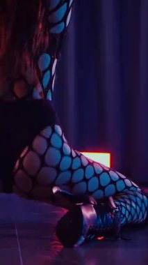 Dikey. Dans eden örgü giysili seksi kadın. Bir dansçı kız karanlık bir salonda erotik bir şekilde dans eder. Kıvranma, yüksek topuklar, striptiz, baştan çıkarıcı bir dans. Atmosferik renkli ışık, kulüp, sahne.