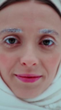 Dikey. Krem rengi ceketli gülümseyen bir kadın kış ormanının ortasında çimlerin üzerinde, karlı bir arazide duruyor. Kış harikalar diyarı; donmuş ağaçların arasında duran bir figür. Kadın portresi, yürü. Karlı orman; atkı takmış bir kadın.