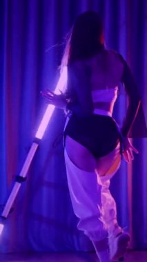 Dikey. Baştan çıkarıcı dans eden seksi kadın. Titreyen bir dansçı, canlı mor ve mavi ışıkla yıkanmış, canlı neon bir üçgenin altında sahnenin enerjisini somutlaştırıyor. Bir sanatçının kadın vücudu.