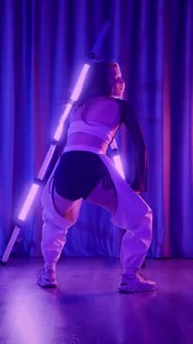 Dikey. Baştan çıkarıcı dans eden seksi kadın. Titreyen bir dansçı, canlı mor ve mavi ışıkla yıkanmış, canlı neon bir üçgenin altında sahnenin enerjisini somutlaştırıyor. Bir sanatçının kadın vücudu.