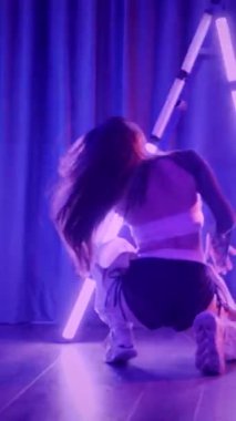 Dikey. Baştan çıkarıcı dans eden seksi kadın. Titreyen bir dansçı, canlı mor ve mavi ışıkla yıkanmış, canlı neon bir üçgenin altında sahnenin enerjisini somutlaştırıyor. Bir sanatçının kadın vücudu.