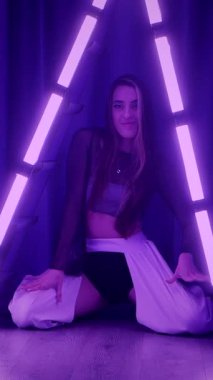 Dikey. Baştan çıkarıcı dans eden seksi kadın. Titreyen bir dansçı, canlı mor ve mavi ışıkla yıkanmış, canlı neon bir üçgenin altında sahnenin enerjisini somutlaştırıyor. Bir sanatçının kadın vücudu.