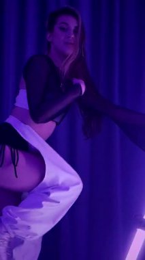 Dikey. Baştan çıkarıcı dans eden seksi kadın. Titreyen bir dansçı, canlı mor ve mavi ışıkla yıkanmış, canlı neon bir üçgenin altında sahnenin enerjisini somutlaştırıyor. Bir sanatçının kadın vücudu.