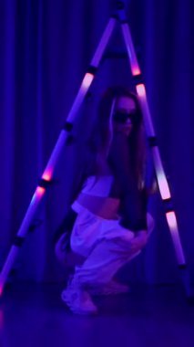 Dikey. Baştan çıkarıcı dans eden seksi kadın. Titreyen bir dansçı, canlı mor ve mavi ışıkla yıkanmış, canlı neon bir üçgenin altında sahnenin enerjisini somutlaştırıyor. Bir sanatçının kadın vücudu.