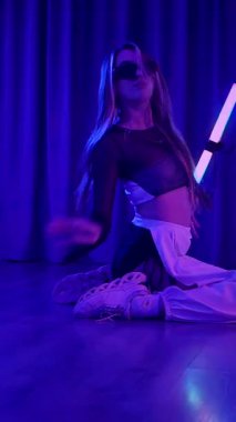 Dikey. Baştan çıkarıcı dans eden seksi kadın. Titreyen bir dansçı, canlı mor ve mavi ışıkla yıkanmış, canlı neon bir üçgenin altında sahnenin enerjisini somutlaştırıyor. Bir sanatçının kadın vücudu.