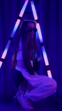 Dikey. Baştan çıkarıcı dans eden seksi kadın. Titreyen bir dansçı, canlı mor ve mavi ışıkla yıkanmış, canlı neon bir üçgenin altında sahnenin enerjisini somutlaştırıyor. Bir sanatçının kadın vücudu.