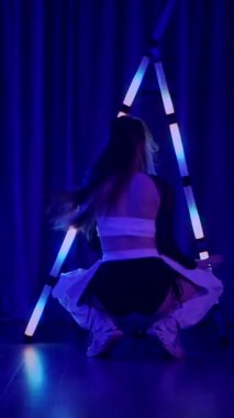 Dikey. Baştan çıkarıcı dans eden seksi kadın. Titreyen bir dansçı, canlı mor ve mavi ışıkla yıkanmış, canlı neon bir üçgenin altında sahnenin enerjisini somutlaştırıyor. Bir sanatçının kadın vücudu.