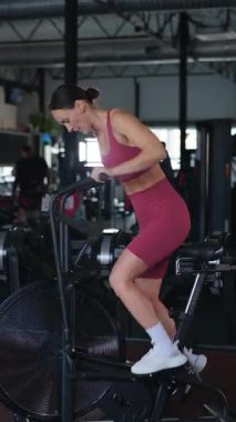 Dikey. Spor salonunda spor bisikletine binen bir kadın. Spor tişörtlü ve şortlu bir kadın aktif olarak antrenman yapıyor, vücudunun harika bir heykelini yaptırıyor..