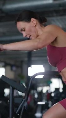 Dikey. Spor salonunda spor bisikletine binen bir kadın. Spor tişörtlü ve şortlu bir kadın aktif olarak antrenman yapıyor, vücudunun harika bir heykelini yaptırıyor..