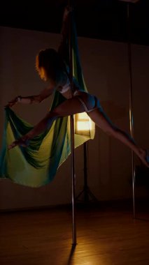 Dikey. Bir kadın ve Teal ipeği bir direğin etrafında dans ediyorlar, büyüleyici bir görüntü. Direk dansı. Karanlıkta asılı kalan bir dansçının zarafeti ortaya çıkar. Bir sanatçının sanatı, stüdyo ışığıyla aydınlatılır..