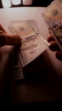 Dikey. Yüz dolarlık banknotların yakın plan görüntüsü. Karmaşık ayrıntıları ince aydınlatılmış. Eller dikkatlice sayıyor, 100 dolarlık banknotlar, yumuşak, sıcak bir ışıkta yıkanmış..