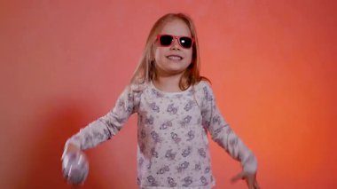 Kırmızı güneş gözlüklü, pijamalı komik kız elinde gümüş bir mikrofon, şarkı söylüyor, dans ediyor ve sıcak turuncu bir fonda poz veriyor. Parlak bir gülümseme ve parlak bir mikrofonla sahneye çıkmaya hazır bir çocuk pop yıldızı..