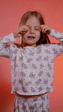 Dikey. Peri desenli pijamalı genç kız geniş bir alanda esniyor ve sıcak bir mercan arka planına yaslanıyor. Çocuğun ağzı esnemiş, uzun sarı saçlar ve açık renkli uyku kıyafetleri sergiliyor..