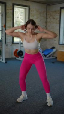 Dikey. Beyaz bluzlu ve pembe taytlı bir kadın spor salonunda spor yapıyor, pilates yapıyor ve esniyor. Bir kız spor salonunda spor yapıyor..