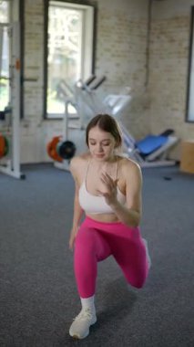 Dikey. Beyaz bluzlu ve pembe taytlı bir kadın spor salonunda spor yapıyor, pilates yapıyor ve esniyor. Bir kız spor salonunda spor yapıyor..