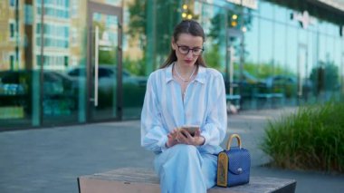 Gözlüklü bir iş kadını sokakta bankta otururken telefonla konuşuyor. Zarif ve başarılı bir kadın akıllı telefondan canlı bir iş görüşmesi yapıyor..