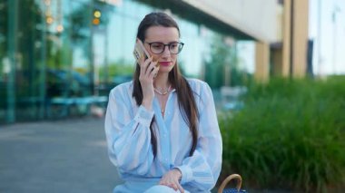 Gözlüklü bir iş kadını sokakta bankta otururken telefonla konuşuyor. Zarif ve başarılı bir kadın akıllı telefondan canlı bir iş görüşmesi yapıyor..