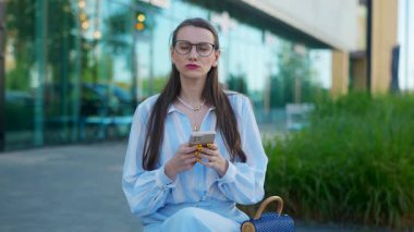 Gözlüklü bir iş kadını sokakta bankta otururken telefonla konuşuyor. Zarif ve başarılı bir kadın akıllı telefondan canlı bir iş görüşmesi yapıyor..