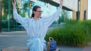 İş kadını tablet üzerinde çalışıyor ve sokakta bir bankta otururken eğleniyor. Genç ve zarif bir kadın dizüstü bilgisayarını elinde tutuyor ve mutlu bir şekilde gülüyor ve ellerini sallıyor..