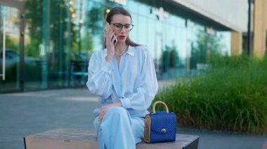 Gözlüklü bir iş kadını sokakta bankta otururken telefonla konuşuyor. Zarif ve başarılı bir kadın akıllı telefondan canlı bir iş görüşmesi yapıyor..