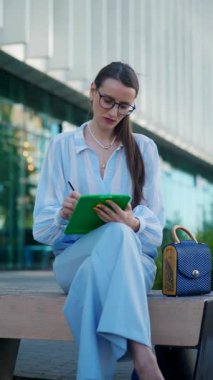 Dikey. Sokaktaki bir bankta otururken iş kadını tablet üzerinde çalışıyor. Zarif genç bayan şehrin ortasında tablet üzerine yoğunlaşarak yazı yazıyor..