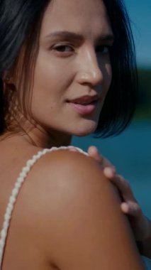 Vertical. A woman, bathed in sunlight, stands by the water's edge, her expression serene. A woman in a delicate cream-colored two-piece swimsuit, poised by the tranquil water. Creamy silk bikini top. Female model posing on the beach.