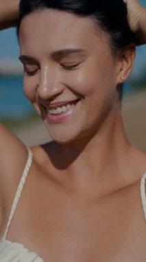 Vertical. A woman, bathed in sunlight, stands by the water's edge, her expression serene. A woman in a delicate cream-colored two-piece swimsuit, poised by the tranquil water. Creamy silk bikini top. Female model posing on the beach.