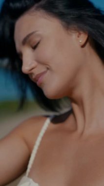 Vertical. A woman, bathed in sunlight, stands by the water's edge, her expression serene. A woman in a delicate cream-colored two-piece swimsuit, poised by the tranquil water. Creamy silk bikini top. Female model posing on the beach.