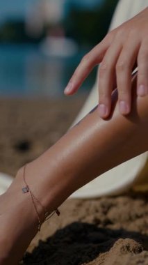 Vertical. A woman, bathed in sunlight, stands by the water's edge, her expression serene. A woman in a delicate cream-colored two-piece swimsuit, poised by the tranquil water. Creamy silk bikini top. Female model posing on the beach.