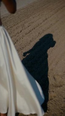 Vertical. A woman, bathed in sunlight, stands by the water's edge, her expression serene. A woman in a delicate cream-colored two-piece swimsuit, poised by the tranquil water. Creamy silk bikini top. Female model posing on the beach.
