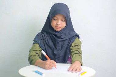 Masada otururken renkli kalemlerle çizen sevimli küçük kız. Güzel Asyalı Müslüman kız beyaz kağıda resim çiziyor.