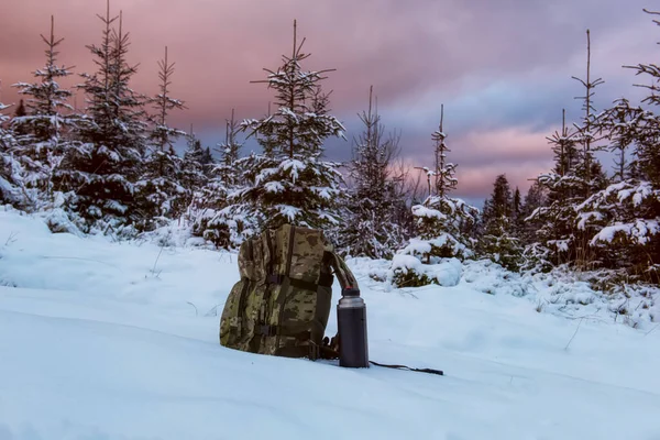 A backpack in military coloring and a thermos on a winter landscape in the forest.