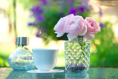 A white cup and delicate pink roses on a table in the garden. Resting place outside in the garden.