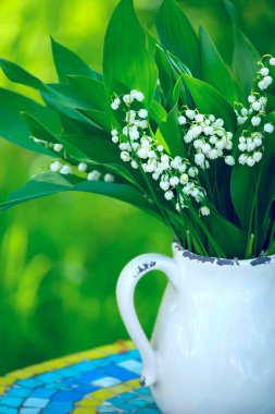 Bir buket bahar çiçeği, yeşil bir bahçenin arka planında seramik bir sürahi içinde vadideki zambaklar..