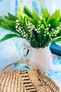 Bahar çiçekleri seramik bir sürahi içinde vadi zambakları ve hasır bir çanta, yaz tatili atmosferi..