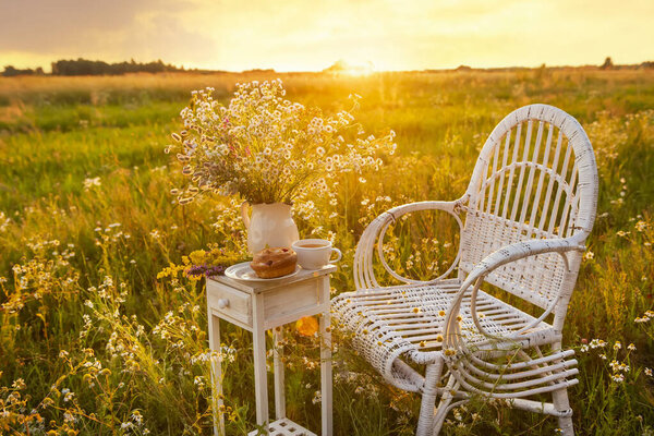 Vintage white furniture table and chair in a field at sunrise or sunset with a cup of tea, a bun and a bouquet of wildflowers. The concept of summer outdoor recreation, relaxation and enjoyment of the moment.