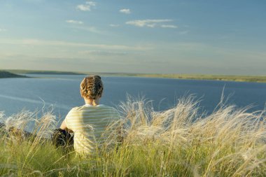 Nehrin kıyısındaki bir uçurumda çiçek açan otların arasında oturan bir kız. Arkadan bir fotoğraf. Açık hava eğlencesi ve yürüyüş..