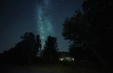Ağaçlar ve samanyolu arasında küçük bir köy evi.